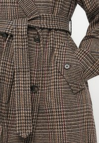 Brown houndstooth coat with a belted waist, large black buttons, side pockets, and a textured woven fabric featuring maroon accents.