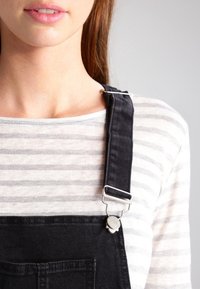 Black denim overalls with silver hardware, featuring wide straps and a large front pocket, worn over a light gray-striped shirt.