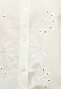 Λευκό ύφασμα με κεντητά λουλουδένια σχέδια που περιλαμβάνουν μικρές οπές και κάθετη λωρίδα με κουμπιά από διάφανο υλικό.