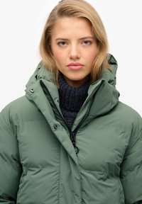 Grüne gepolsterte Jacke mit hohem Kragen und Kapuze, ausgestattet mit einem Reißverschluss und Druckknopfverschluss, getragen über einem dunklen Strickpullover.