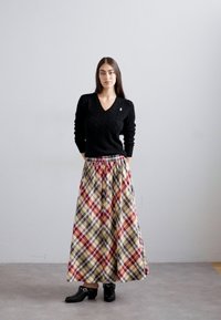 Egy nő áll, kezét a háta mögött tartva, fekete pulóvert, hosszú, színes kockás szoknyát és fekete bokacipőt viselve egy szürke padlón.