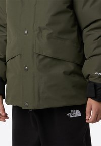 Grønn vattert jakke med teksturert overflate, med metalliske trykknapper og svarte detaljer. Sorte joggebukser med hvit logo.