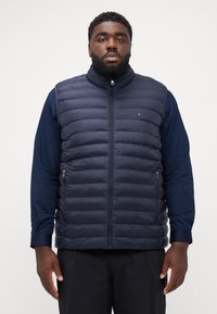 Gilet matelassé bleu marine avec un col haut, fermeture éclair sur le devant et poches latérales. Présente une texture matelassée horizontale et un petit logo sur la poitrine.