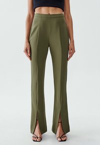 Maßgeschneiderte olivgrüne Hose mit hoher Taille, Bügelfalten vorne und seitlichen Schlitzen, aus einem glatten Stoff gefertigt. In Sicht sind schwarze Riemchensandalen.