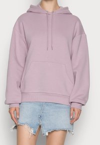 Sudadera con capucha de color púrpura claro, con un bolsillo delantero y cordones. Cuenta con mangas amplias y puños de canalé. Combinada con una falda corta de mezclilla.