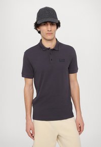 Polo oscuro gris con mangas cortas y un corte ajustado, que presenta un sutil logo de EA7. Combinado con un pantalón beige claro y una gorra gris.
