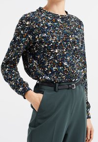 Donna che indossa una blusa lunga a maniche lunghe con fantasia floreale scura, infilata in pantaloni verdi scuri a vita alta con una cintura nera, mano in tasca.