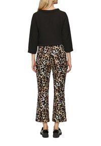 Schwarzes strukturiertes, kurz geschnittenes Oberteil mit Dreiviertelärmeln, kombiniert mit tierisch bedruckten, weit ausgestellten Hosen in beigen, braunen und schwarzen Farbtönen. Schwarze Schuhe.