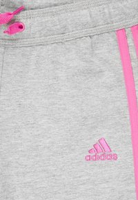 Šedé tepláky s růžovým stažením a růžovými bočními pruhy. Na levé přední straně je vyšité logo Adidas. Měkká textura, příležitostný design.
