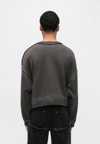 Σκούρο γκρι βαμβακερό φούτερ με κομψό cropped σχέδιο, ακατέργαστες άκρες και δύο διακοσμητικά μεταλλικά κουμπώματα στο πίσω �μέρος του λαιμού. Φορεμένο με φθαρμένο μαύρο τζιν.