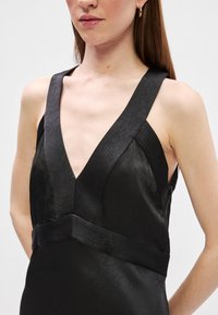 Čierne šaty s hlbokým výstrihom do V, širokými ramienkami, textúrovanou tkaninou a priliehavým pásom s ozdobným šitím. Podrobný dizajn zdôrazňuje jednoduchosť.