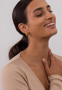Boucles d'oreilles en or avec une finition lisse et brillante. Le modèle porte un top beige ajusté et côtelé, coiffé d'une queue de cheval basse.