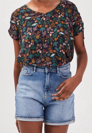 Personne portant un chemisier à manches courtes à motif floral foncé, rentré dans un short en denim bleu clair à ourlet roulé, avec une main près de la poche du short.
