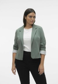 Světlá zelená blazer s vyhrnutými rukávy p�řes bílou košili, doplněná černými kalhotami. Hladká látka, strukturovaný design a zapínání na jeden knoflík.