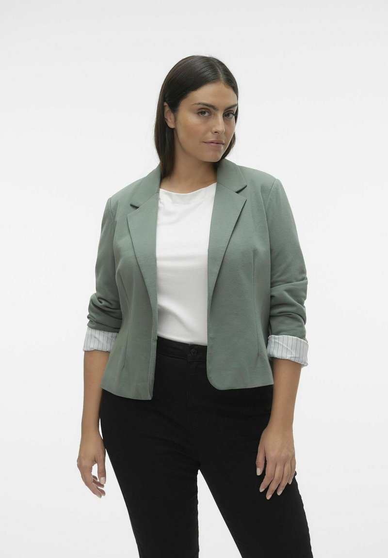Světlá zelená blazer s vyhrnutými rukávy přes bílou košili, doplněná černými kalhotami. Hladká látka, strukturovaný design a zapínání na jeden knoflík.