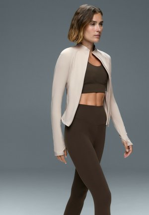 Vrouw met kort haar die een lichtbeige jas met rits draagt over een donkerbruine sportbeha en hooggetailleerde legging, lopend tegen een grijze achtergrond.