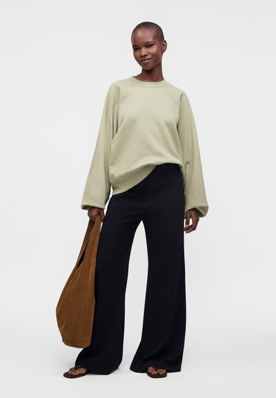 Une personne souriante se tient debout, tenant un grand sac cabas marron, vêtue d'un pull ample vert clair, d'un large pantalon noir et de sandales, sur un fond uni.