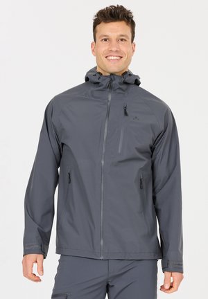 Homme souriant portant une veste imperméable grise à capuche avec poches zippées et pantalon assorti, sur un fond uni.