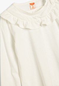 Langärmliges, cremefarbenes Baumwollhemd mit gerafftem Ausschnitt und einem Etikett, das die Größe 104-110 cm angibt. Weiche, glatte Textur.