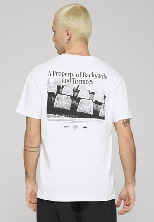 Mann med kort blondt hår sett bakfra, iført en hvit T-skjorte med svart grafikk og teksten "A Property of Backyards and Terraces."