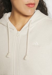 Detailní záběr na osobu v krémové Adidas mikině na zip s kapucí a viditelným vyšitým logem na hrudi.