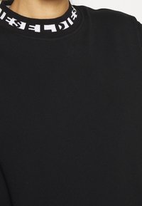 Sudadera negra con un cuello alto y una banda con logo en blanco contrastante. Textura suave y diseño ajustado.