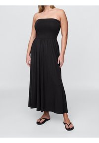 Svart strapless maxiklänning med en figurnära bodice, flödig kjol och mjukt tyg. Buren med svarta flip-flops mot en neutral bakgrund.