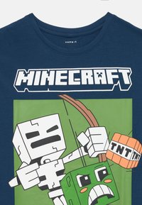 Tamsiai mėlynas medvilnės marškinėliai su "Minecraft" logotipu ir grafika, kurioje skeleto figūra laiko TNT šalia žalio veikėjo su rimtu veidu.