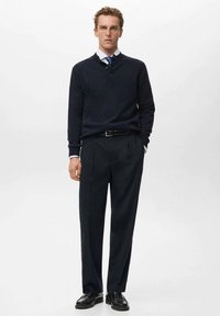 Maglione lavorato a maglia navy con collo alto e dettagli in bottone, abbinato a pantaloni scuri sartoriali e scarpe nere per un look formale.