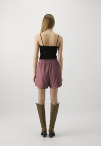 Černý top na ramínka, volné růžové šortky s texturovaným vzorem a hnědé kloboukové boty po kolena s mírným podpatkem. Pohled zezadu na tento outfit.