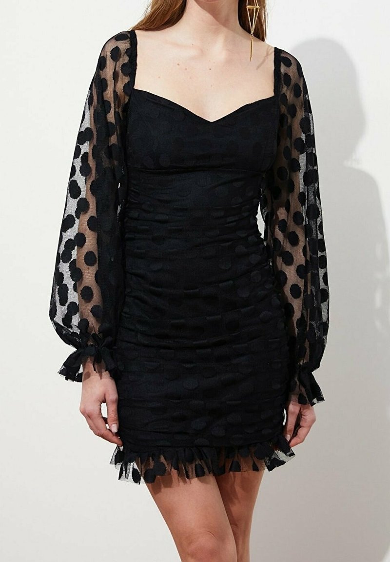 Femme portant une robe noire ajustée avec des manches longues transparentes à pois et un ourlet à volants, debout devant un fond clair.