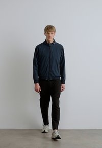 Tumesinine bomber-jakk lukuga ja elastsete mansettidega, yhdistatud mustade kitsaste pükstega ja valgete tossudega, seistes valge tausta ees.