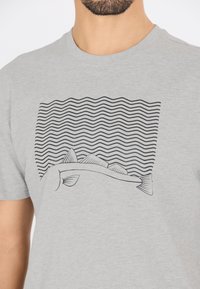 Harmaa puuvillapaita, jossa on musta viivapiirros kalasta, jossa on evät aalto-kuvioisen taustan edessä. Yksinkertainen, graafinen muotoilu.