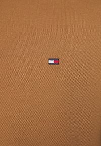 Tissu tricoté marron avec un petit logo brodé aux accents bleu, blanc et rouge, positionné sur le côté gauche.