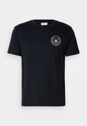 Mørkeblå bomull t-skjorte med en rund grafikk-patch med røde og hvite detaljer. Klassisk rund hals og korte ermer, glatt tekstur.