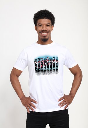 Un homme souriant se tient les mains sur les hanches, portant un T-shirt blanc avec des personnages des Avengers en costumes rouges et noirs.