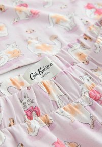 Stof met speelse katten in verschillende posities, op een roze achtergrond en met een zichtbaar Cath Kidston-label nabij de geplooide naden.