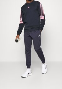 A person walks while wearing a dark sweater with pink stripes and matching joggers, holding a water bottle in one hand.