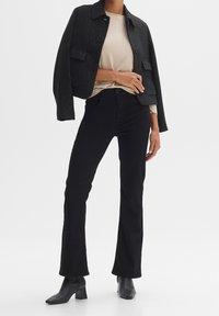 Veste noire texturée avec col, poches avant ; haut beige côtelé ; pantalon noir évasé ; bottines noires à talons.