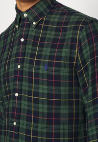 Green plaid shirt made of fabric with button-down collar. Features navy, yellow, and red grid pattern, with a small embroidered logo.