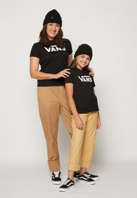 Dos personas llevan camisetas negras de Vans y gorros. Combinan las partes superiores con pantalones beige y zapatillas negras con detalles blancos.