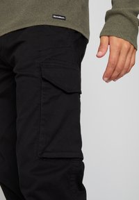 Oliwkowy sweter dzianinowy z prążkowanymi mankietami, zestawiony z czarnymi spodniami cargo z bocznymi kieszeniami i gładką fakturą.