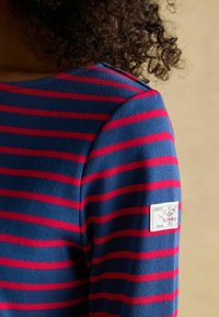 Sinine ja punane triibuline pika varrukaga särk, mis on valmistatud ribakangast, vasakul õlal logo plaastriga. Disaini lähivõte.