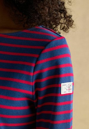 Langärmliges Shirt mit blauen und roten Streifen aus geripptem Stoff, mit einem Logo-Patch auf der linken Schulter. Nahaufnahme des Designs.