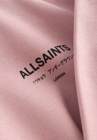 Różowa tkanina z czarnym napisem „ALLSAINTS”, japońskimi znakami i napisem „LONDON” nadrukowanymi na powierzchni.