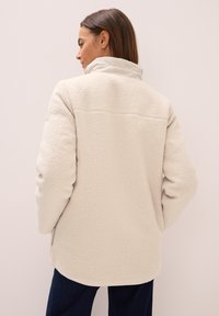 Veste en polaire de couleur crème clair avec une surface texturée, col montant et ourlet légèrement arrondi. Comprend un empiècement au dos et des poches latérales.