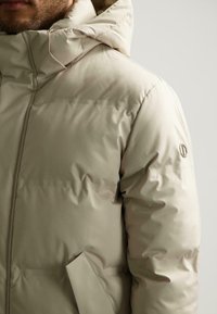 Beige pufferjack met een capuchon, voorzien van een gladde buitenstof, een geborduurd patroon en een zijzak met minimale branding op de mouw.