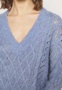 Maglione lavorato a maglia di colore azzurro chiaro con scollatura a V, caratterizzato da un motivo a rombi, dettagli a trecce e un design a punto aperto, che conferisce un aspetto texture.