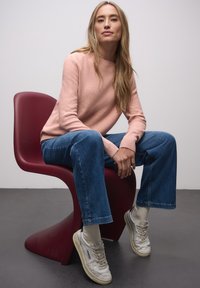 Jasnoró�żowy dziergany sweter, niebieskie dżinsy o szerokich nogawkach i szare tenisówki. Kobieta siedzi na burgundowym krześle na tle neutralnym.