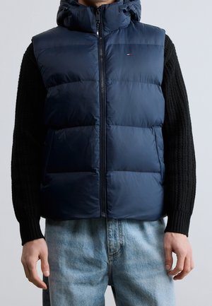 Mężczyzna ubrany w granatową pikowaną bezrękawnik puchową na czarnym dzianinowym swetrze i jasnoniebieskich dżinsach, stojący na jednolitym tle.